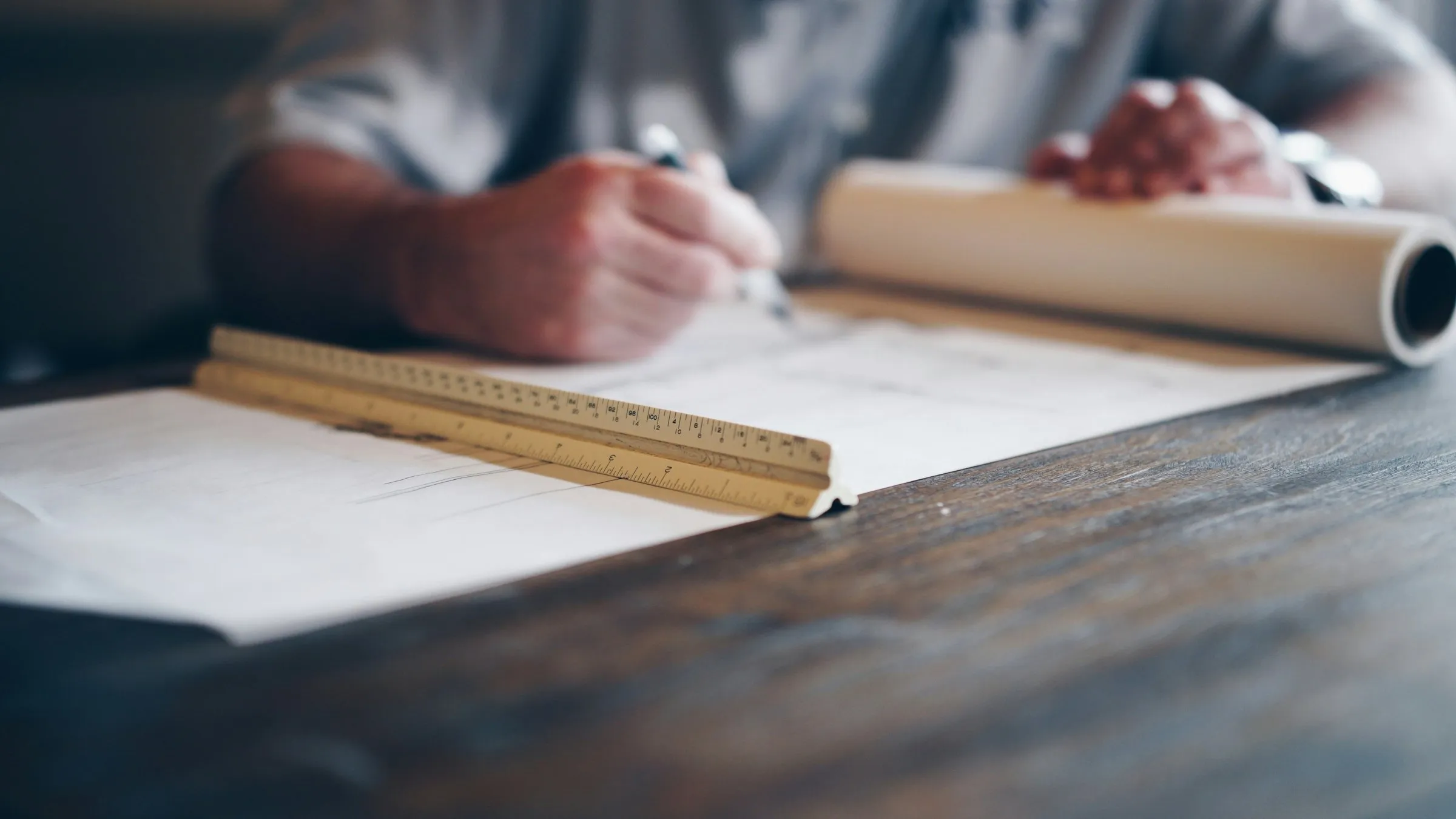 Hands drafting a plan on a dark wooden table, next to a ruler and a roll of paper