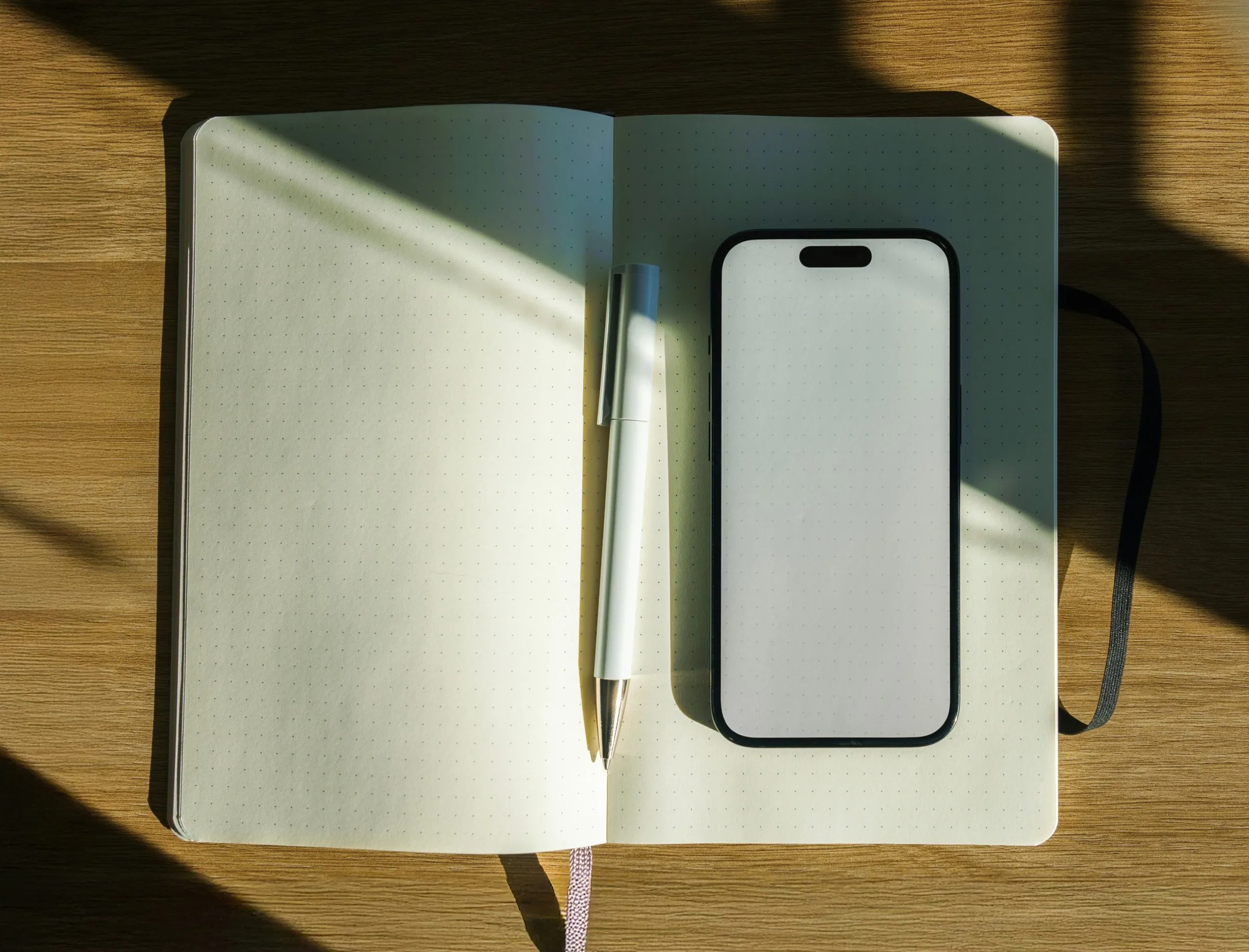A smartphone and a notebook with a pen on a wooden table in sunlight
