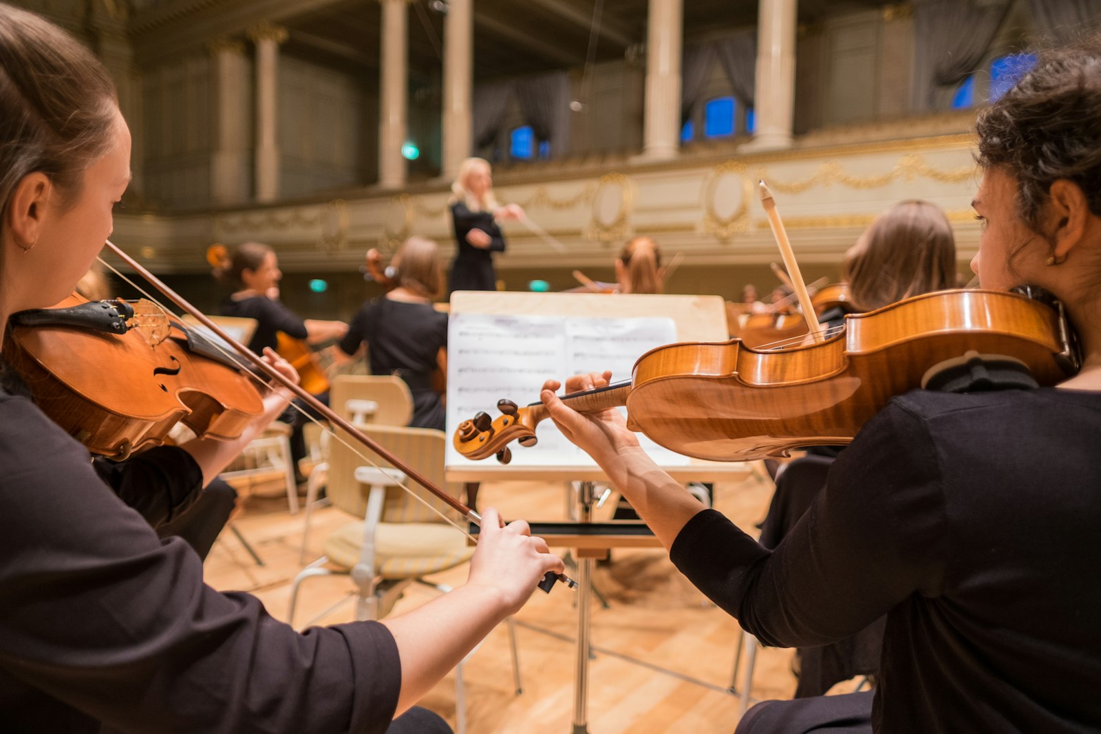 Zwei Geigerinnen im Orchester spielen im Zusammenspiel, im Hintergrund die Dirigentin